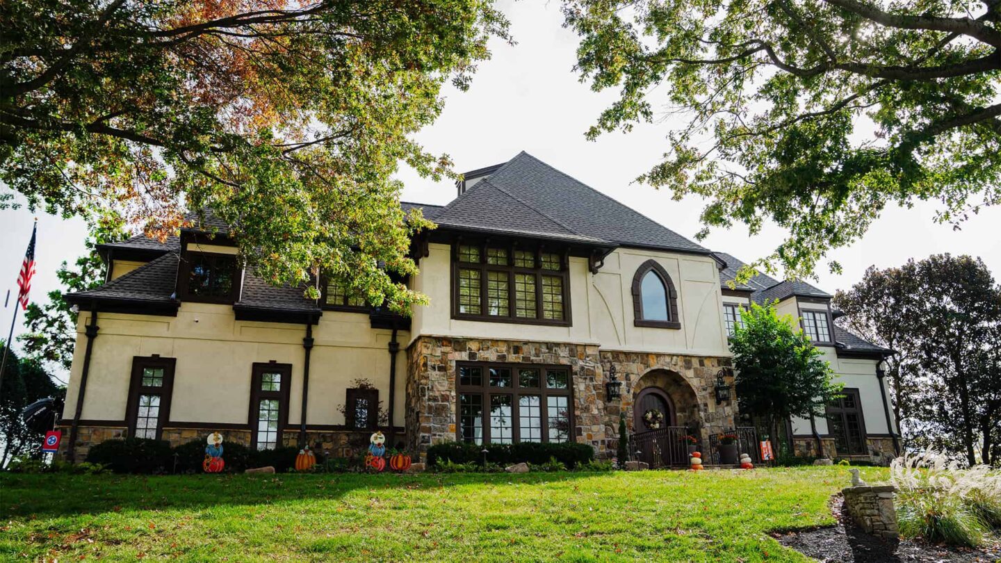 A large two-story house with stone and stucco exterior, featuring metal roofing, surrounded by trees and a grassy lawn, decorated with fall and Halloween items.