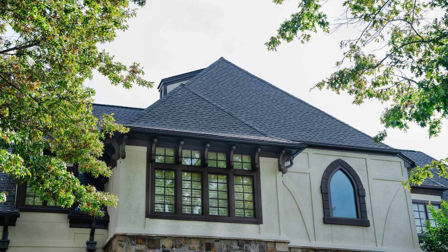 A large house with cream-colored stucco walls, dark roof shingles by Barnett Roofing, and large windows, surrounded by green leafy trees.