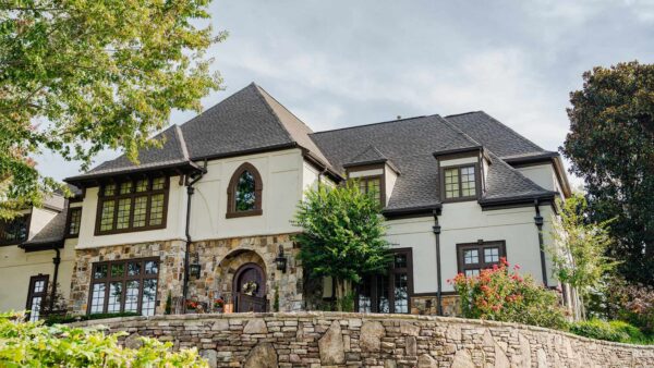 Large two-story stone and stucco house with a dark roof by Barnett Roofing, numerous windows, and landscaping, viewed from behind a stone retaining wall under a partly cloudy sky.