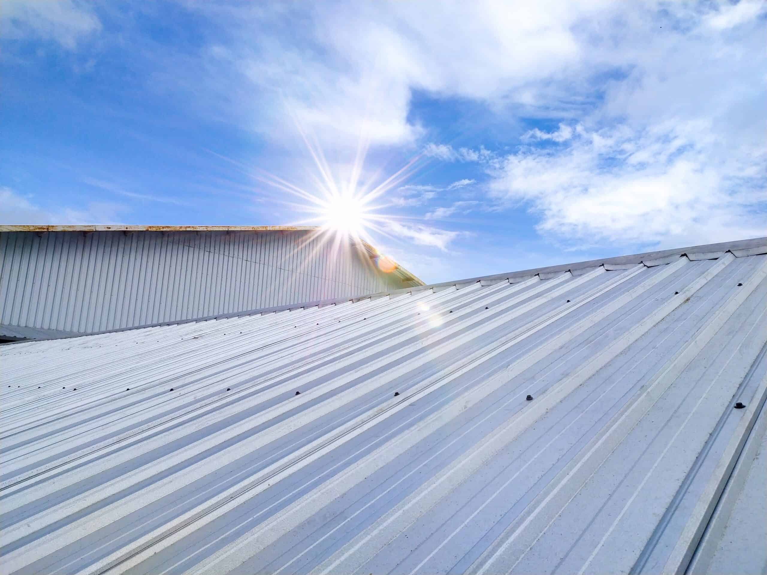 Sun shines above the edge of a corrugated metal roofing, with a blue sky and clouds in the background—a classic touch you can expect from Barnett Roofing.