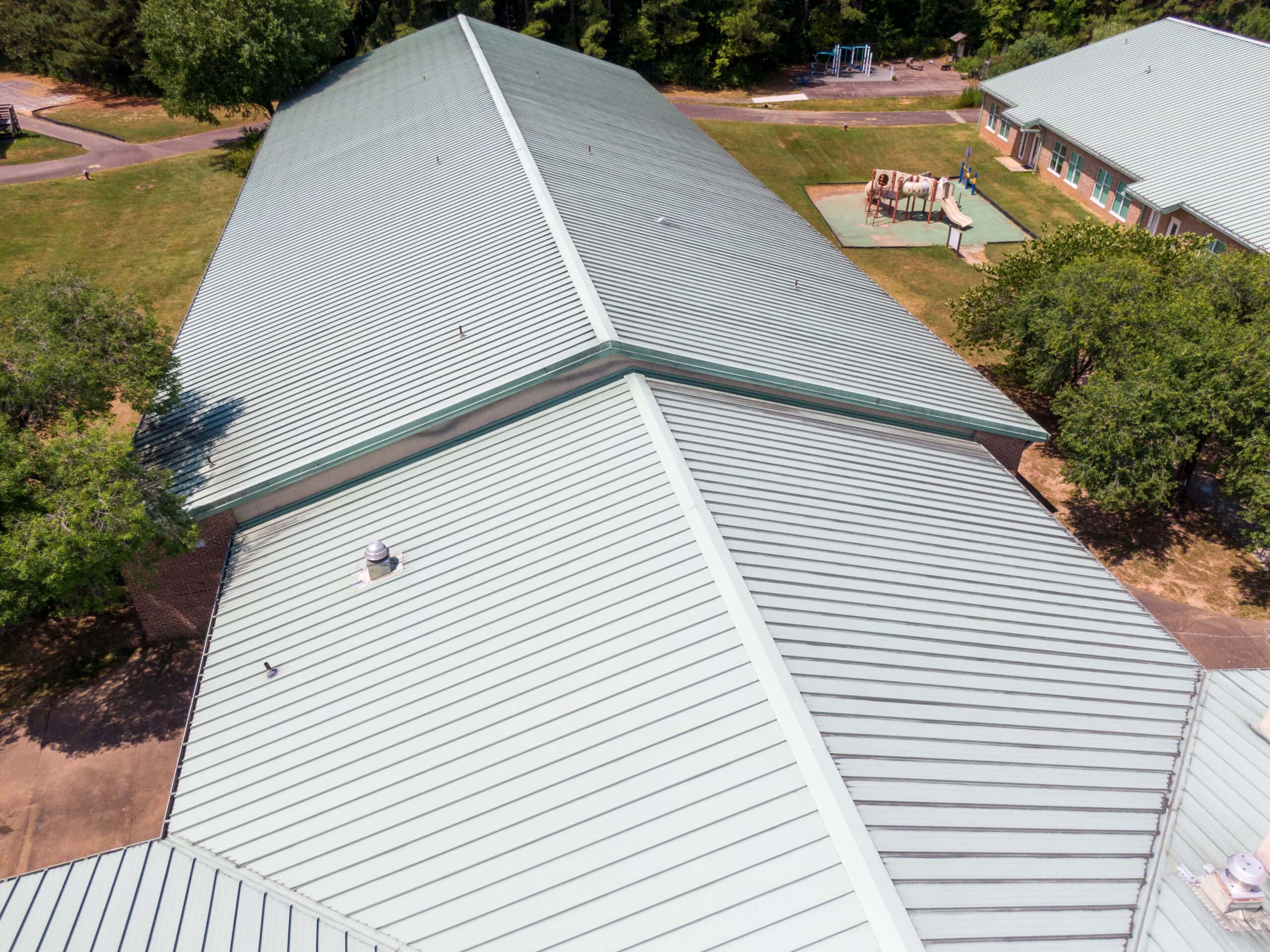 Aerial view of a large commercial roofing project featuring a metal roof on a building, surrounded by grass, trees, and walkways, with an adjacent playground visible in the background.