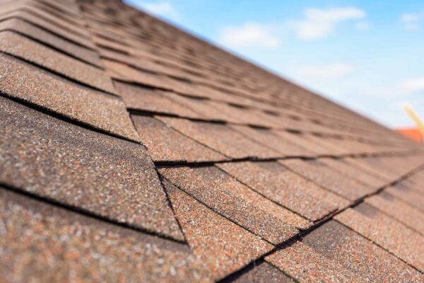 Close-up view of brown asphalt shingles on a sloped roof under a blue sky with scattered clouds, showcasing Barnett Roofing’s expertise in both residential and commercial roofing solutions.