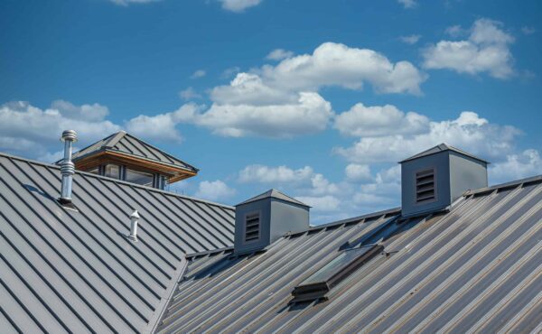Gray metal rooftops with vents and a skylight, part of quality metal roofing, are shown under a blue sky with scattered white clouds.