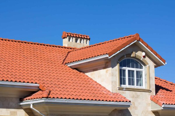 House roof with red clay tiles, a chimney, and a decorative dormer window showcases expert residential roofing by Barnett Roofing against a beige stone exterior under a clear blue sky.