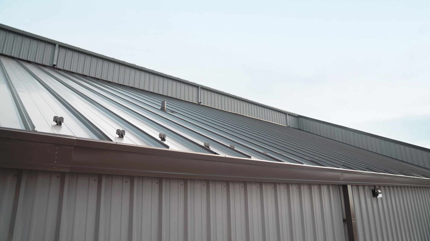 A close-up view of a sloped metal roof and siding on a building by Barnett Roofing, featuring vertical seams and fasteners under a clear sky.
