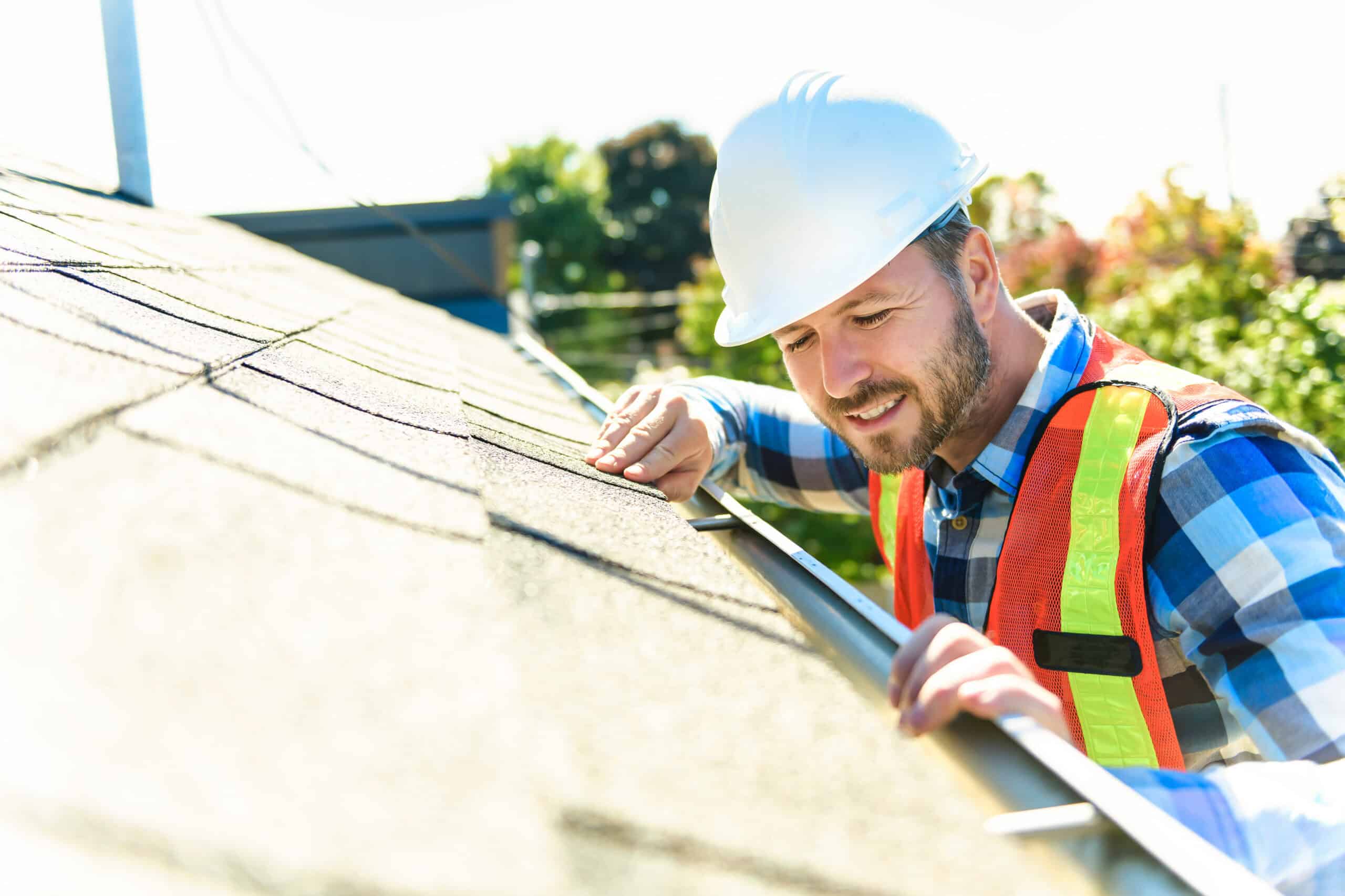 A roofer in safety gear inspecting roof shingles. He wears a white hard hat, blue plaid shirt, and high-visibility vest. The worker is leaning over the edge of a roof, carefully examining the shingles with his hand. The background shows a sunny day with greenery visible.