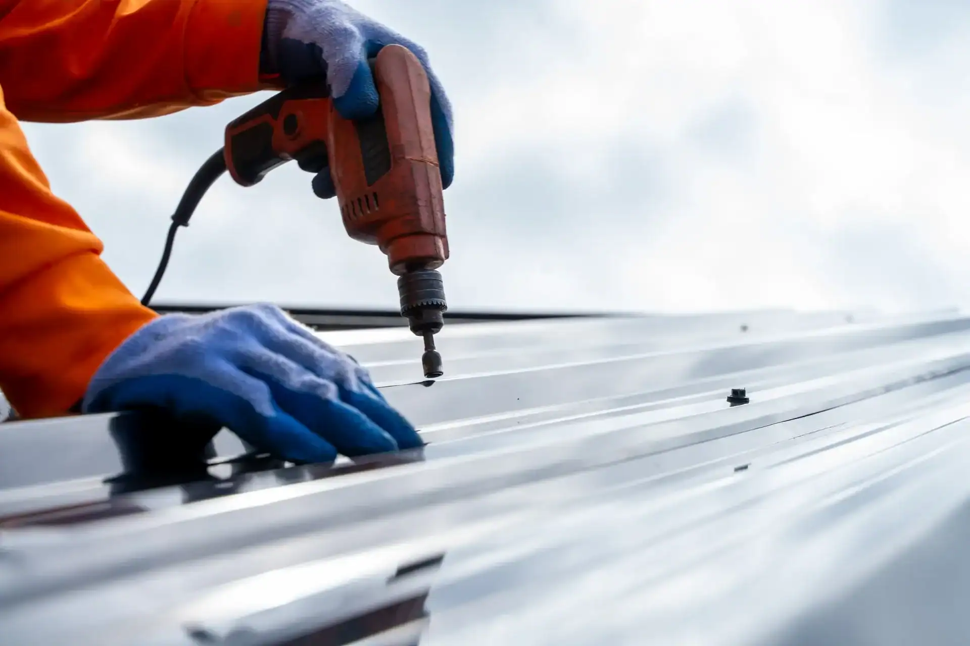 A person wearing gloves and an orange sleeve uses a power drill to fasten screws into a metal roof panel, showcasing Barnett Roofing’s commitment to quality residential roofing.