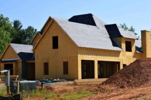 A house under construction with exposed plywood walls and a roof partially covered with underlayment, surrounded by dirt and building materials, showcases the initial stages of roofing installation.