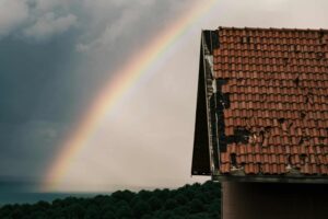 A rainbow appears in the cloudy sky above treetops, with a partially damaged tiled roof and hints of metal roofing in the foreground.