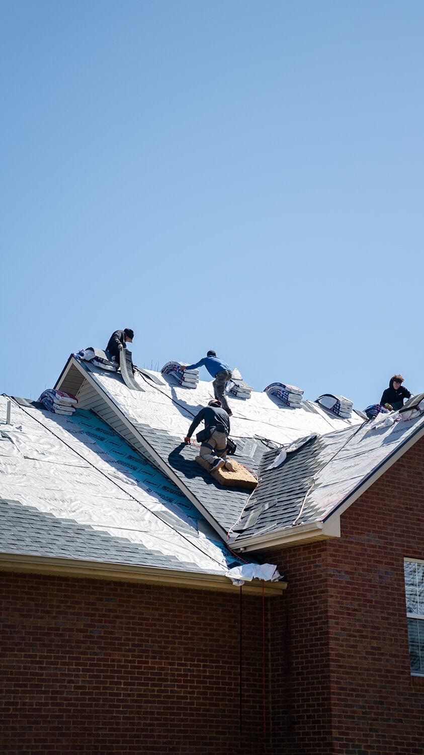 Several workers from Barnett Roofing are installing metal roofing materials on the roof of a brick house under a clear blue sky.