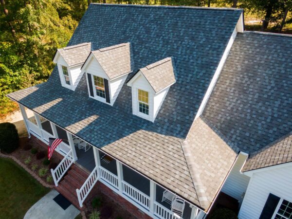 Aerial view of a house with gray asphalt shingle roofing, white dormer windows, and a front porch with an American flag. Trees and lawn are visible in the background.