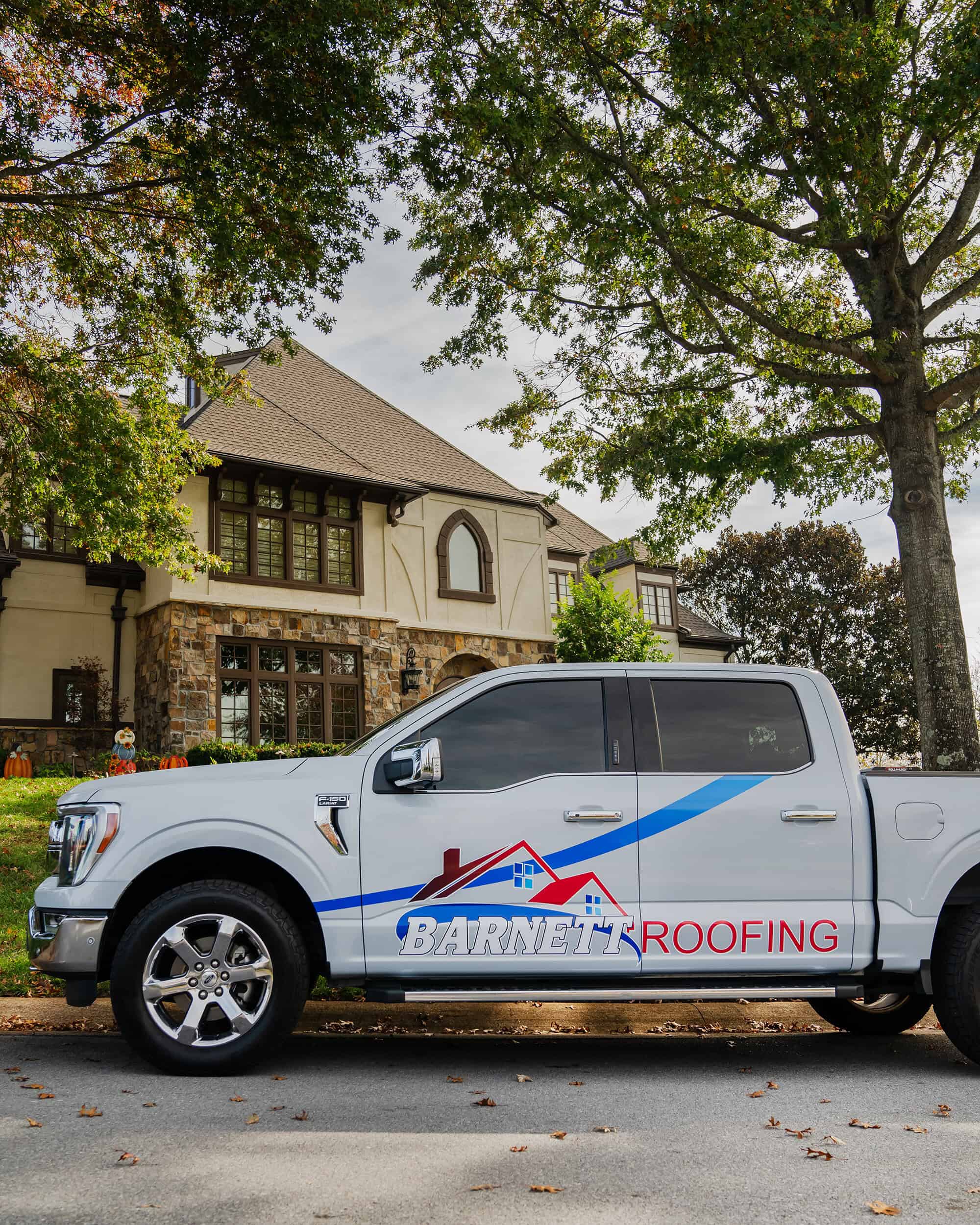 A white pickup truck with "Barnett Roofing" branding is parked on a suburban street in front of a large house with stone and stucco exterior, highlighting their expertise in residential roofing.