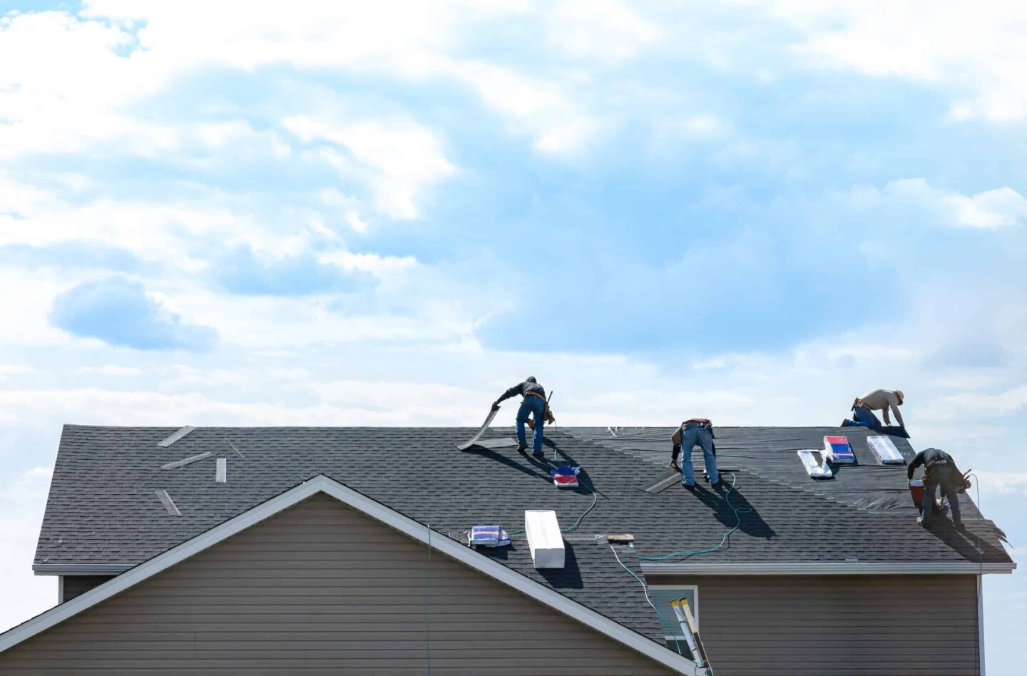 Four workers from Barnett Roofing are installing or repairing shingles on the roof of a house under a partly cloudy sky. Roofing materials and equipment are scattered around them.